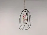 Et hængende ornament med sorte trådløkker, hvide perler og et lille blomstermotiv med pink og orange blomster i midten.