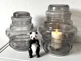 To riflede glasvaser, hvoraf den ene holder et tændt hvidt lys, står ved siden af en lille sort-hvid pandafigur på en hvid flade mod en lys væg.