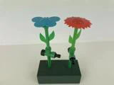 To havesprinkler i plast formet som blomster, en blå og en rød, med grønne stilke og blade, monteret på en grøn rektangulær base mod en hvid baggrund.