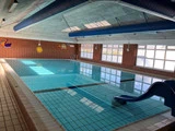 Indendørs swimmingpool med klart blåt vand, klinkegulv, røde murstensvægge, store vinduer, vægkunst og en lille blå rutsjebane ned i poolen.