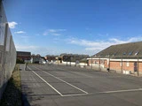 En indhegnet udendørs tennisbane uden spillere, omgivet af murstensbygninger og huse under en klar blå himmel.