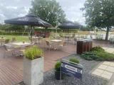 En udendørs terrasse med flere borde og stole, store sorte parasoller, planter og et skilt med teksten »INDGANG«. I baggrunden ses træer og en overskyet himmel.