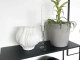 En hvid tekstureret vase, et tændt hvidt lys i en glasholder og en grøn potteplante står på en sort hylde mod en hvid væg.