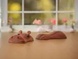 To flodhesteskulpturer af træ, den ene viser hovedet med øjne og ører, og den anden viser ryggen, på et træbord med et sløret vindue og blomster i baggrunden.