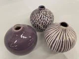 Tre runde, lilla keramikvaser med smalle åbninger, hver med et forskelligt struktureret mønster, udstillet på en lys overflade.