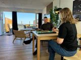 To personer sidder ved et træbord og spiller backgammon i en lys, moderne stue med store vinduer og udsigt til en balkon.