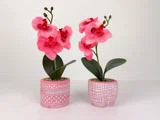 To lyserøde kunstige orkidéer i mønstrede lyserøde og hvide potter, placeret side om side på en almindelig hvid flade mod en hvid baggrund.