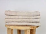 Fire pænt foldede beige håndklæder stablet på en rund træskammel mod en almindelig hvid struktureret væg.