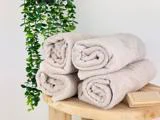 Fire pænt sammenrullede beige håndklæder er stablet på en træplade ved siden af en lille børste, og i baggrunden hænger en grøn bladplante.