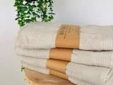 En stak beige badehåndklæder med brune papiretiketter ligger på en træplade med grønne hængende planteblade i baggrunden.