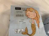 En lyseblå serviet har en illustreret smilende havfrue med langt orange hår og en guldhale ved siden af teksten LITTLE MERMAID.