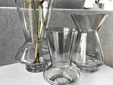 Tre tomme klare glasvaser med moderne geometriske former står på en hvid flade; i den ene vase er der et par høje blomsterstilke. I baggrunden er der grå, strukturerede møbler.