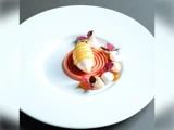 En dessert på tallerken med en central hvirvel af pink sauce, en quenelle toppet med gul sauce, klatter af fløde, orange gelé og spiselige lilla blade på en hvid tallerken.