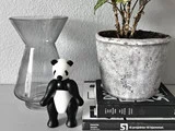 En glasvase, en lille pandafigur, en potteplante og stablede bøger er arrangeret på en lysegrå overflade mod en struktureret hvid væg.