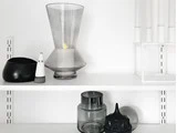 En minimalistisk hylde med dekorative ting: en stor glasvase med et stearinlys, en sort skål, en lille spøgelsesfigur, stablede glasbeholdere og hvide stearinlys i en holder.