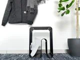 En quiltet sort jakke og et gråt tørklæde hænger på væggen over et moderne sort skostativ med en hvid sneaker; en potteplante står til højre.