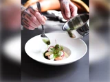 En tatoveret kok hælder grøn sauce fra en gryde over en gourmetret med radiser og krydderurter med en ske på en hvid tallerken.