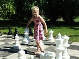 En ung pige i en lyserød kjole spiller skak med store udendørs brikker på et bræt i naturlig størrelse i en solrig park.