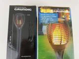 To æsker med Grundig sollysflammer, som hver viser et billede af en fakkelformet udendørs solcellelampe med flammeeffekt. Den ene æske er sort, den anden er blå.
