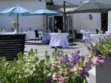 Udendørs terrasse med borde dækket af hvide duge, skygget af blå og grå parasoller; lilla og pink blomster i forgrunden; hvid bygning i baggrunden.