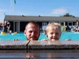 En skægget mand og en ung pige smiler, mens de kigger ud over kanten af en pool med vand og mennesker i baggrunden på en solrig dag.