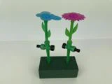 To farverige blomsterformede havesprinkler i plast, en blå og en lilla, er fastgjort til grønne stængler på en grøn rektangulær base.