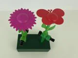 To sprinklere til haveslanger, den ene formet som en pink blomst og den anden som en rød sommerfugl, er monteret på en grøn, rektangulær base.