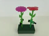 To blomsterformede haveslanger af plast, en lyserød og en rød, er monteret på grønne stængler med stik og står oprejst i en grøn skumblok.
