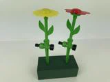 To blomsterformede haveslanger af plast, en gul og en rød, står oprejst i en grøn skumblok mod en almindelig hvid baggrund.