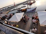 Folk samles på en træbrygge nær en havn med sejlbåde ved solnedgang; nogle sidder under store hvide parasoller, andre står og snakker.