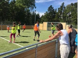 Flere personer iført lyse veste spiller fodbold på en lille udendørs bane, mens nogle få tilskuere ser til bag en træbarriere under en solrig himmel.