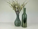 To grønne glasvaser: den ene med hvide kunstige blomster og grene, den anden tom, placeret side om side mod en almindelig lys baggrund.