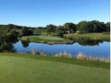 En golfgreen ligger på en ø omgivet af vand, med træer og buske i baggrunden under en klar blå himmel.