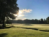 En solbeskinnet golfbane med grønt græs, sandbunker og træer rundt om kanten under en klar blå himmel med nogle skyer.