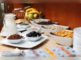 En morgenbuffet med skåle med morgenmadsprodukter, tørret frugt, frø og en kande mælk med en kurv med frisk frugt i baggrunden.