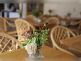 En lille buket blomster i en glasvase står på et træbord i en café med kurvestole og flere borde i den slørede baggrund.