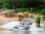 Udendørs restaurantterrasse med borde og stole, dækkeservietter til spisning, potteplanter, grønne planter og dekorative lanterner langs en stenmur.