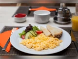En tallerken med røræg, ristet brød og frisk salat med tomat, agurk og salat, sat på et bord med juice, en kop og bestik.