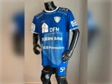 En sort mannequin iført en blå fodboldtrøje med forskellige sponsorlogoer og sorte shorts, der står indendørs foran en ensfarvet baggrund.