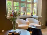 Et hyggeligt opholdsområde med to hvide lænestole ved et vindue, en vase med blomster på et bord, bøger og blødt naturligt lys, der strømmer ind.