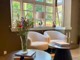 Et hyggeligt siddeområde med to hvide stole ved et vindue, en vase med blomster på et bord, bøger og moderne indretning i et godt oplyst rum.