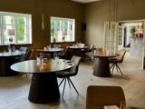 En moderne, elegant restaurant med runde træborde, pænt dækket med glas og bestik, brune stole og store vinduer, der lukker naturligt lys ind.