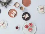 Fem tallerkener med kagestykker, en tallerken med fire æbler, krus med kaffe, en lille skål med frø og en buket blomster arrangeret på en hvid bordplade.