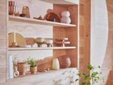 Træhylder indbygget i en lys trævæg rummer dekorative keramikgenstande, skåle, bøger og planter og skaber en minimal, varm og hyggelig æstetik. Sollyset kaster skygger på scenen.