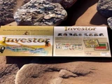 Et brætspil, der hedder Investor, ligger åbent på sten udendørs og viser spilpenge, kort og instruktioner inde i æsken.