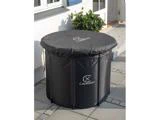 En rund, sort Capablex udendørs opbevaringsbeholder med monteret låg er placeret på en flisebelagt terrasse i nærheden af et hvidt hus og en grøn potteplante.