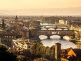 En naturskøn udsigt over Firenze ved solnedgang med Arnofloden, historiske broer og klassisk italiensk arkitektur med bakker i baggrunden.