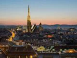 Et bybillede af Wien ved solnedgang med Stephansdom med sit høje spir og farverige tag, omgivet af historiske bygninger og blødt oplyst af aftenhimlen.