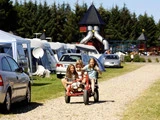 Tre børn kører på en gokart på en grussti på en campingplads med telte, biler og en legeplads med rutsjebaner i baggrunden.