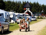 Tre børn kører glade på en firehjulet trækvogn på en grussti på en campingplads, hvor man kan se telte, biler og en legeplads med rutsjebaner i baggrunden.